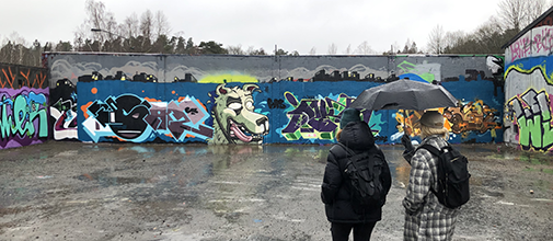 Graffitimålningar på en lång vägg i ett industriområde. Två personer står och tittar på dem.